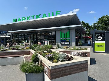 Een zomerse foto van Marktkauf Meppen. Je kunt de ingang zien met het groene logo en bankjes met planten.